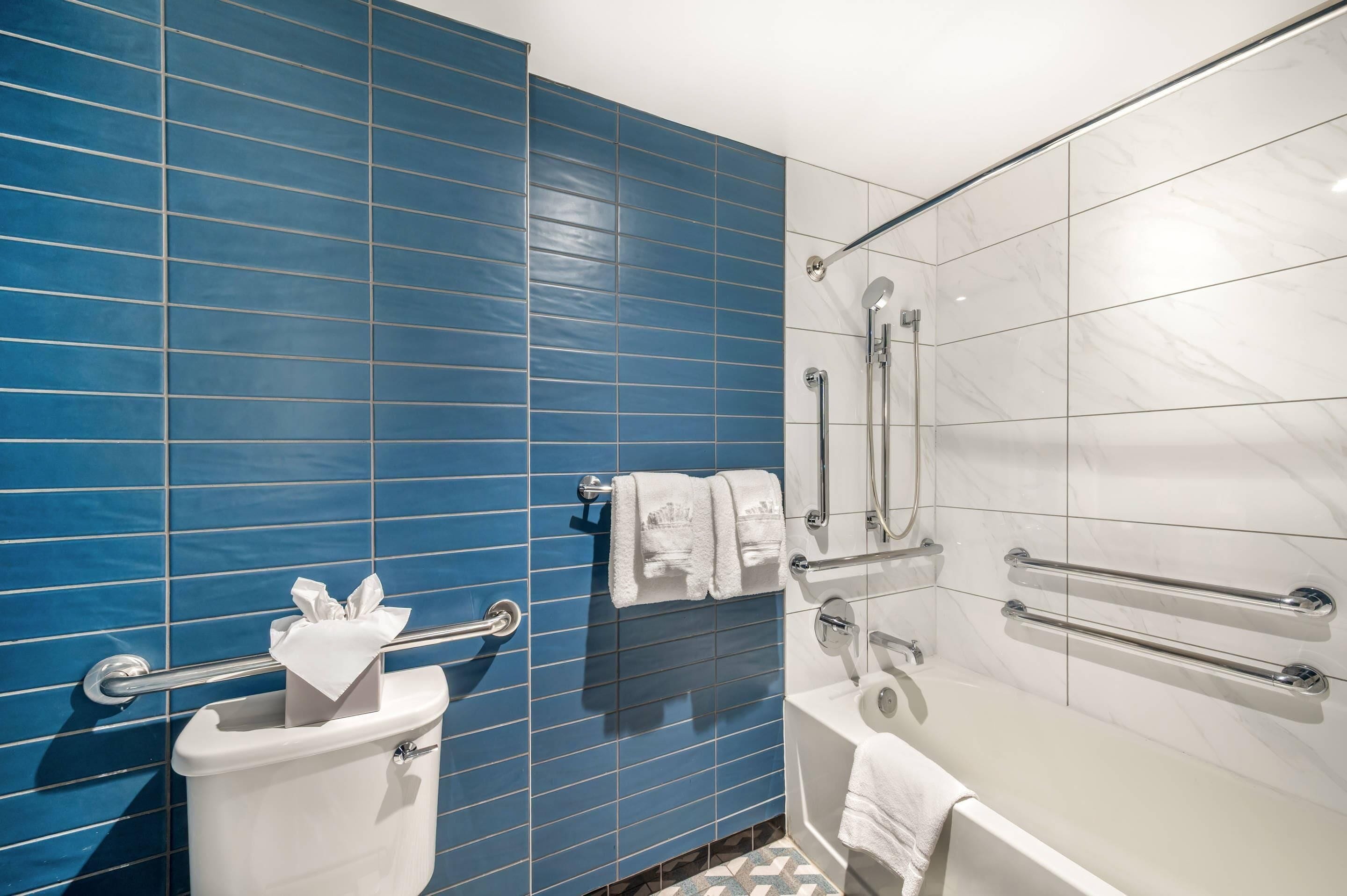 Un baño con paredes de azulejos azules, inodoro blanco, bañera y ducha con barras de apoyo.