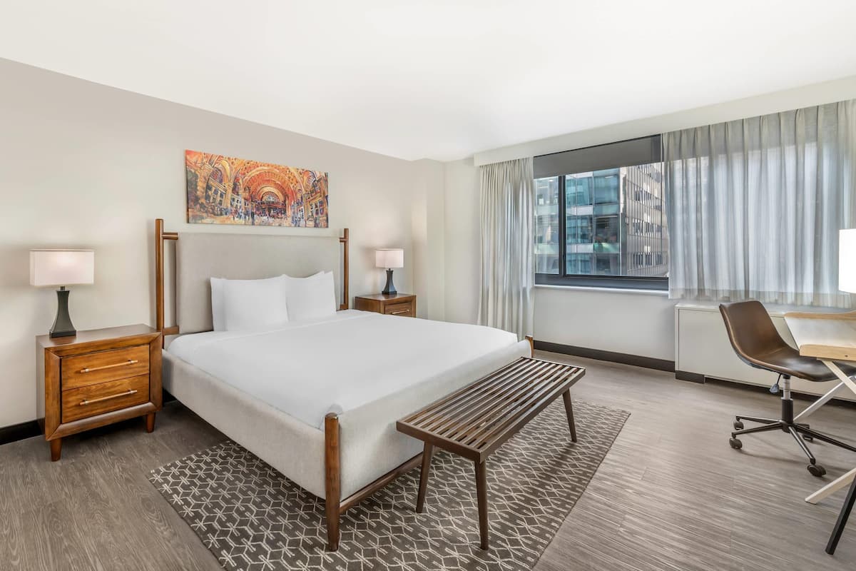 Une chambre d’hôtel comprenant un lit, des tables de chevet, un bureau, une chaise, une fenêtre avec des rideaux et un tableau au mur.