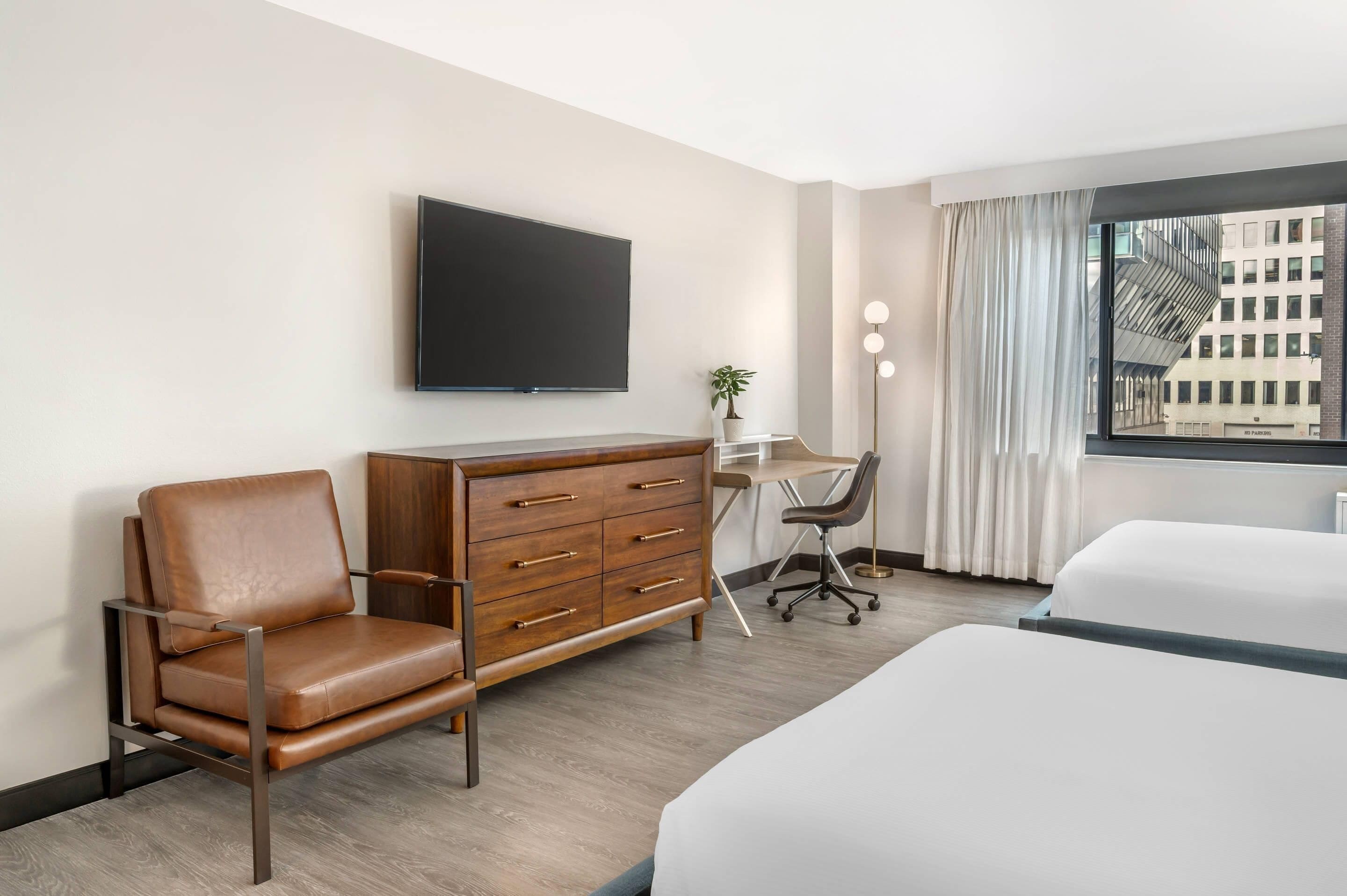 A hotel room with a bed, a desk, a chair, a TV, and a window with a view of buildings.