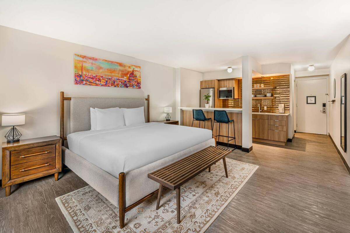 Une chambre d’hôtel moderne avec un grand lit, un banc en bois, un bureau et une kitchenette.