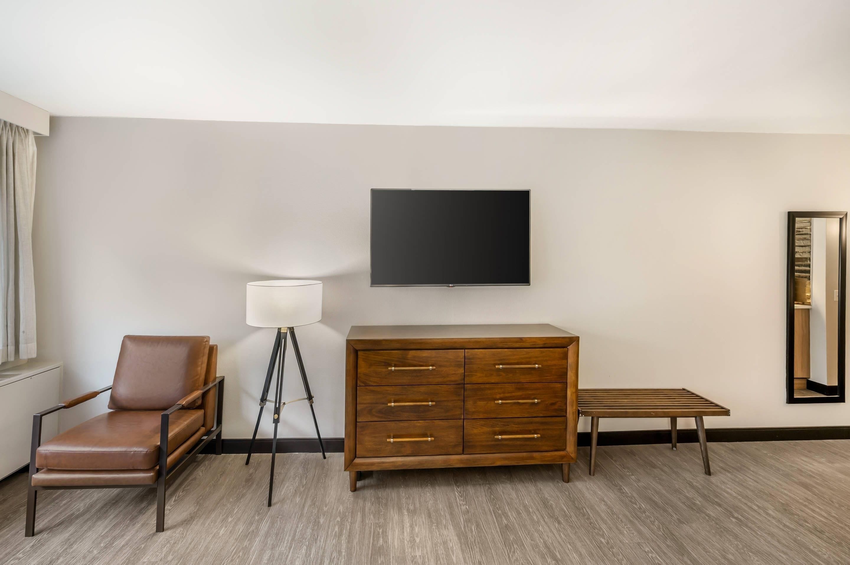 Habitación de hotel con un sillón de cuero marrón, un aparador de madera, una mesita pequeña, una lámpara de pie y un televisor de pantalla plana montado en la pared.