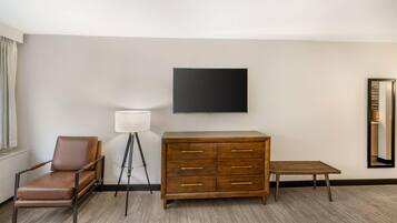 Habitación de hotel con un sillón de cuero marrón, un aparador de madera, una mesita pequeña, una lámpara de pie y un televisor de pantalla plana montado en la pared.