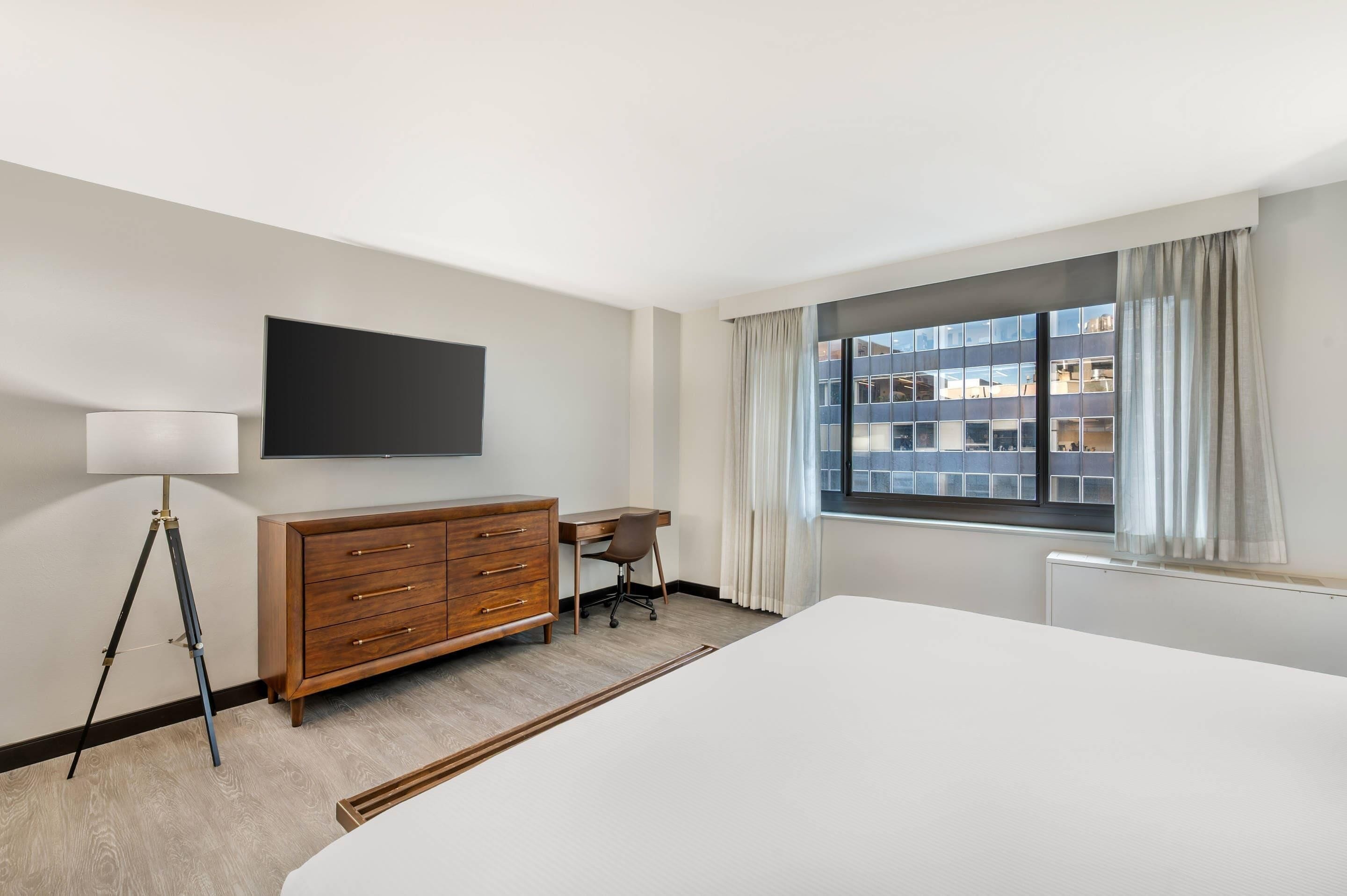 A hotel room with a bed, a desk, a dresser, a lamp, a TV, and a window with curtains.