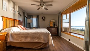 Ein Schlafzimmer mit Bett, Deckenventilator, Kommode, Spiegel und Fenster mit Blick auf den Strand.