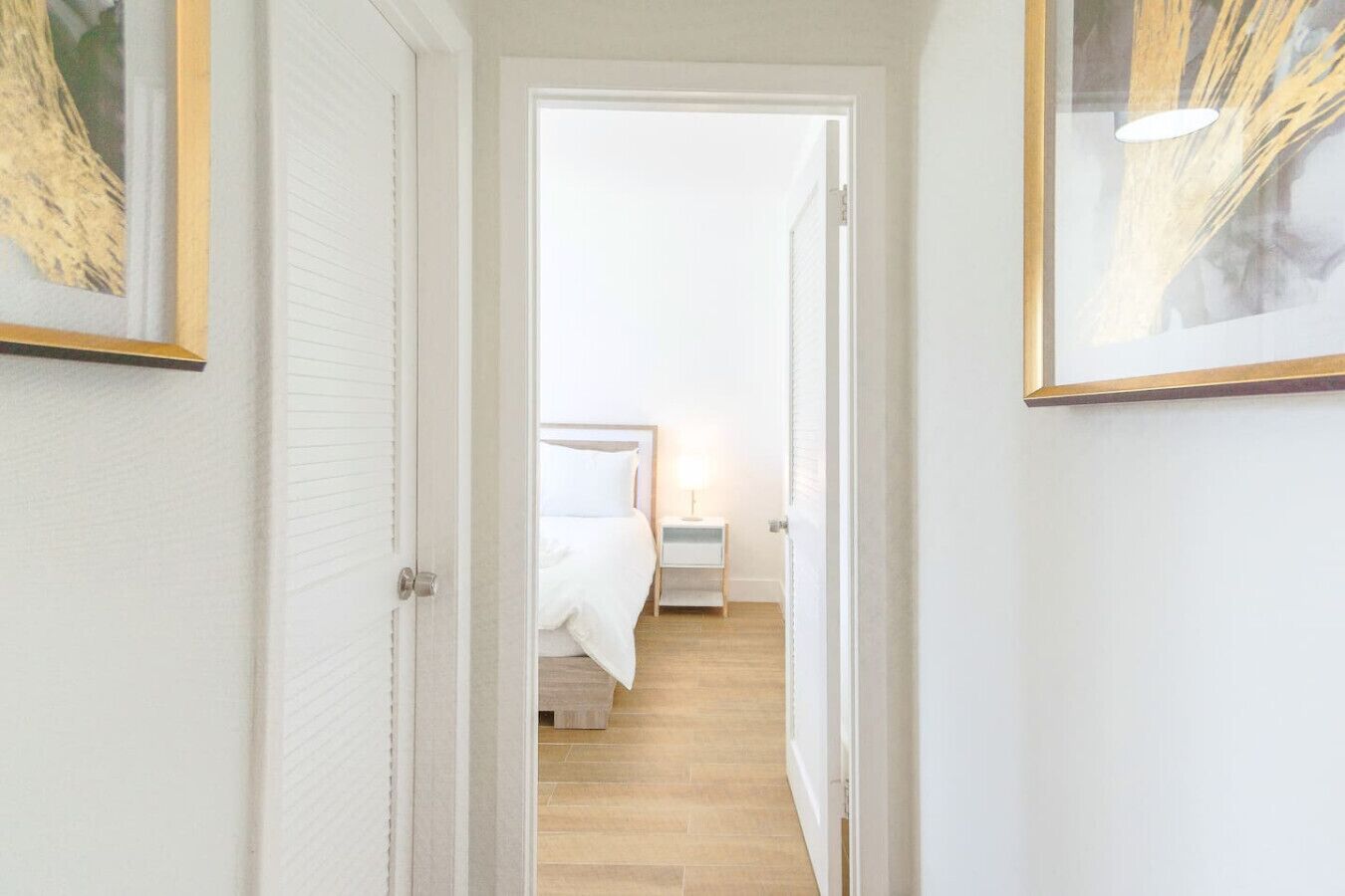 Un couloir étroit mène à une chambre à coucher qui contient un lit, une table de chevet et une lampe.