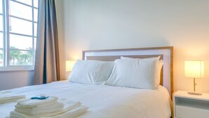 A neatly made bed with white linens and pillows, a stack of folded towels, and two bedside lamps on a nightstand.