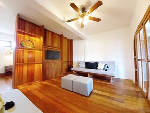 Ein modernes Wohnzimmer mit Holzvertäfelung, Deckenventilator, Flachbildfernseher, einer gepolsterten Couch und einem kleinen weißen Hocker.