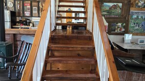 A wooden staircase with a handrail, leading to a room with a kitchen area, wooden cabinets, and various signs and posters on the walls. - Stone Road Log House (Westport)
