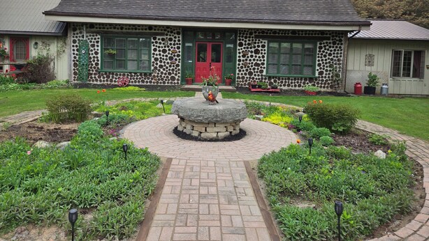 A stone house with a red door, green shutters, and a garden with a circular stone feature in the center. - Stone Road Log House (Westport)