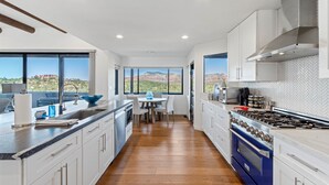 Une cuisine moderne avec des meubles blancs, un poêle bleu et une vue sur les montagnes grâce à de grandes fenêtres.