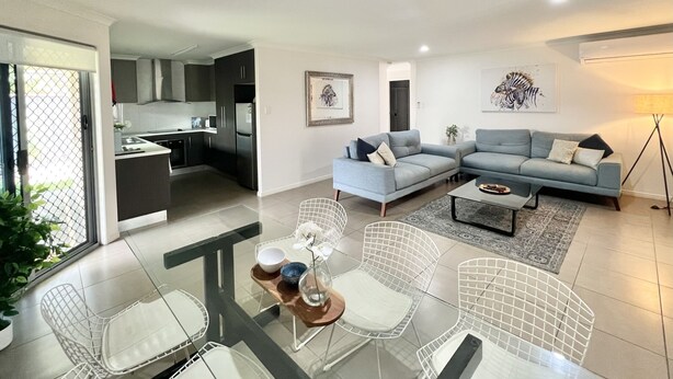 A modern living room with a grey sofa, a dining table with chairs, and a kitchen in the background.