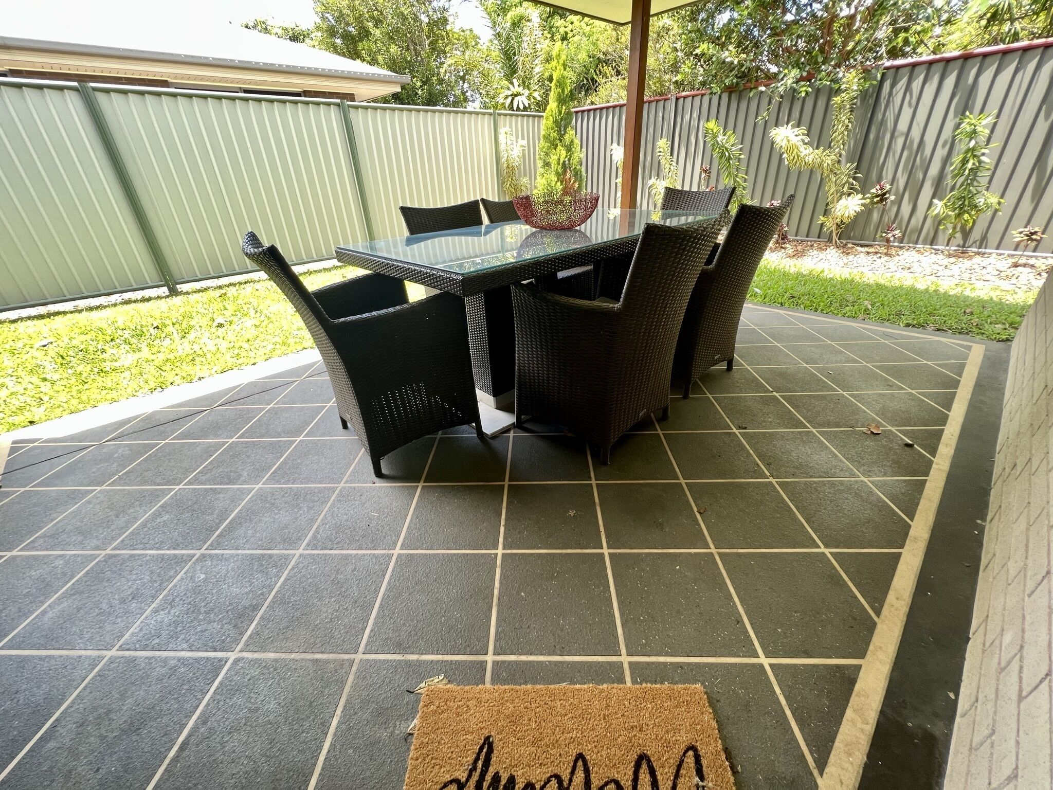 Een terras met een tafel met glazen blad en rieten stoelen, omringd door een groene hekken en een betegelde vloer.