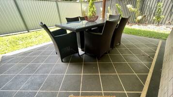 Een terras met een tafel met glazen blad en rieten stoelen, omringd door een groene hekken en een betegelde vloer.