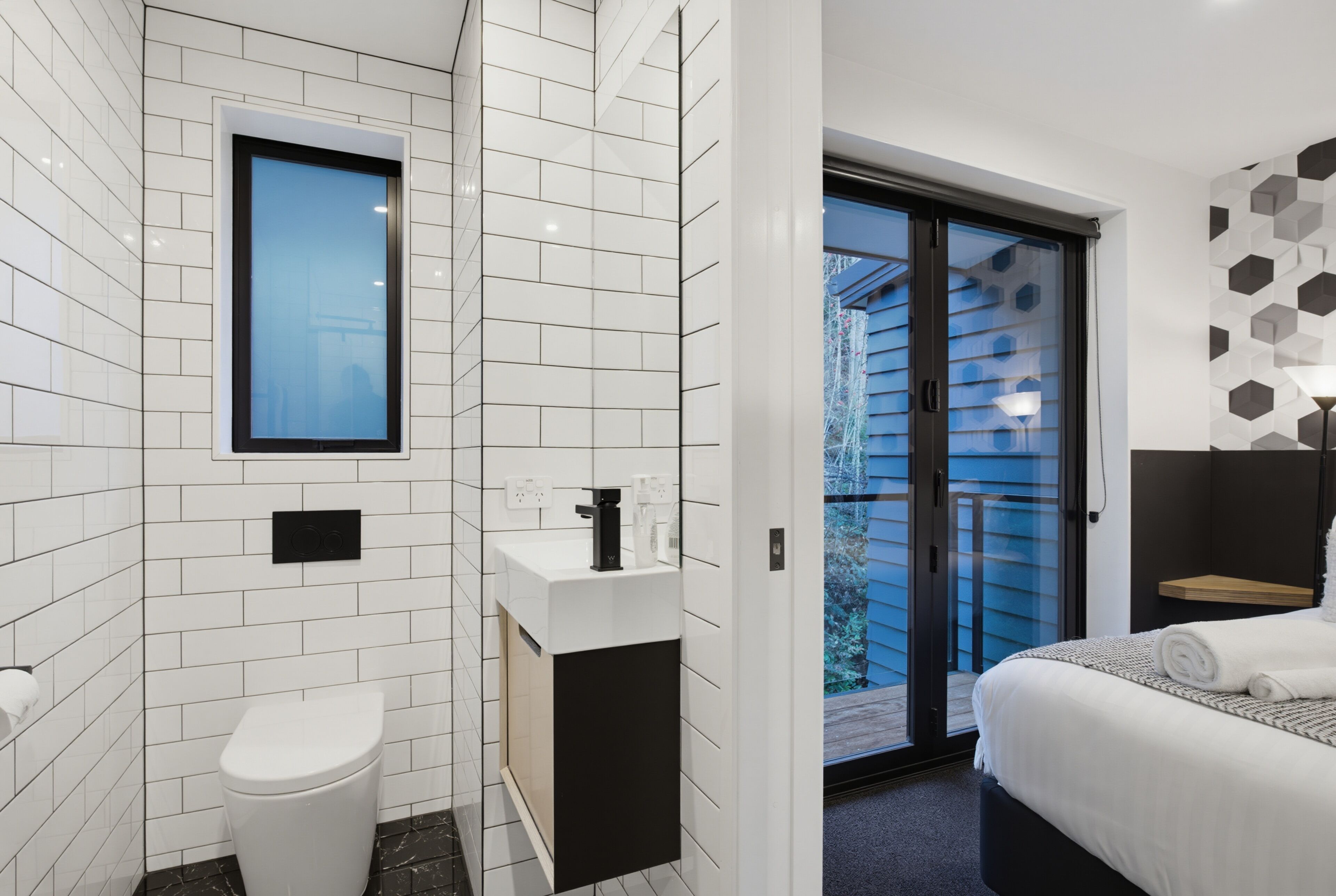 A modern bathroom with a toilet, sink, and shower, a balcony with a view, and a bedroom with a bed and a nightstand.