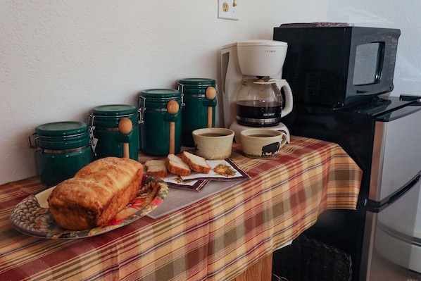 Et køkken med mikrobølgeovn, kaffemaskine og grønne opbevaringsbeholdere på et bord med brød og en underlag.