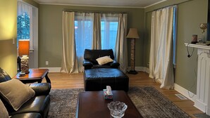 A living room with a leather sofa, a coffee table, and a lamp. There is a window with curtains and a view of the outside.