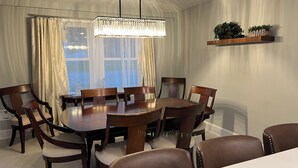 A dining room with a round table, six wooden chairs, a chandelier, and a window with curtains.
