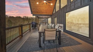 Un espace extérieur abrité, aménagé pour les repas, avec une longue table et des chaises, un ventilateur de plafond et de grandes fenêtres offrant une vue sur le paysage environnant.