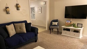 Ein Wohnzimmer mit einem dunkelblauen Sofa, einem weißen Couchtisch, einer Glasvase mit Dekoration, einer Ablage für Fernbedienungen und einem Flachbildfernseher auf einem weißen Schrank.