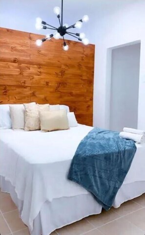 A bedroom with a wooden headboard, a white bed with multiple pillows, a blue blanket, and a modern chandelier.