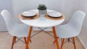 Ein runder, weißer Tisch mit zwei weißen Stühlen, jeweils mit Holzbeinen. Auf dem Tisch stehen zwei weiße Teller, ein kleines Topfpflänzchen und ein Paar Essstäbchen auf einer Holz Untertasse.