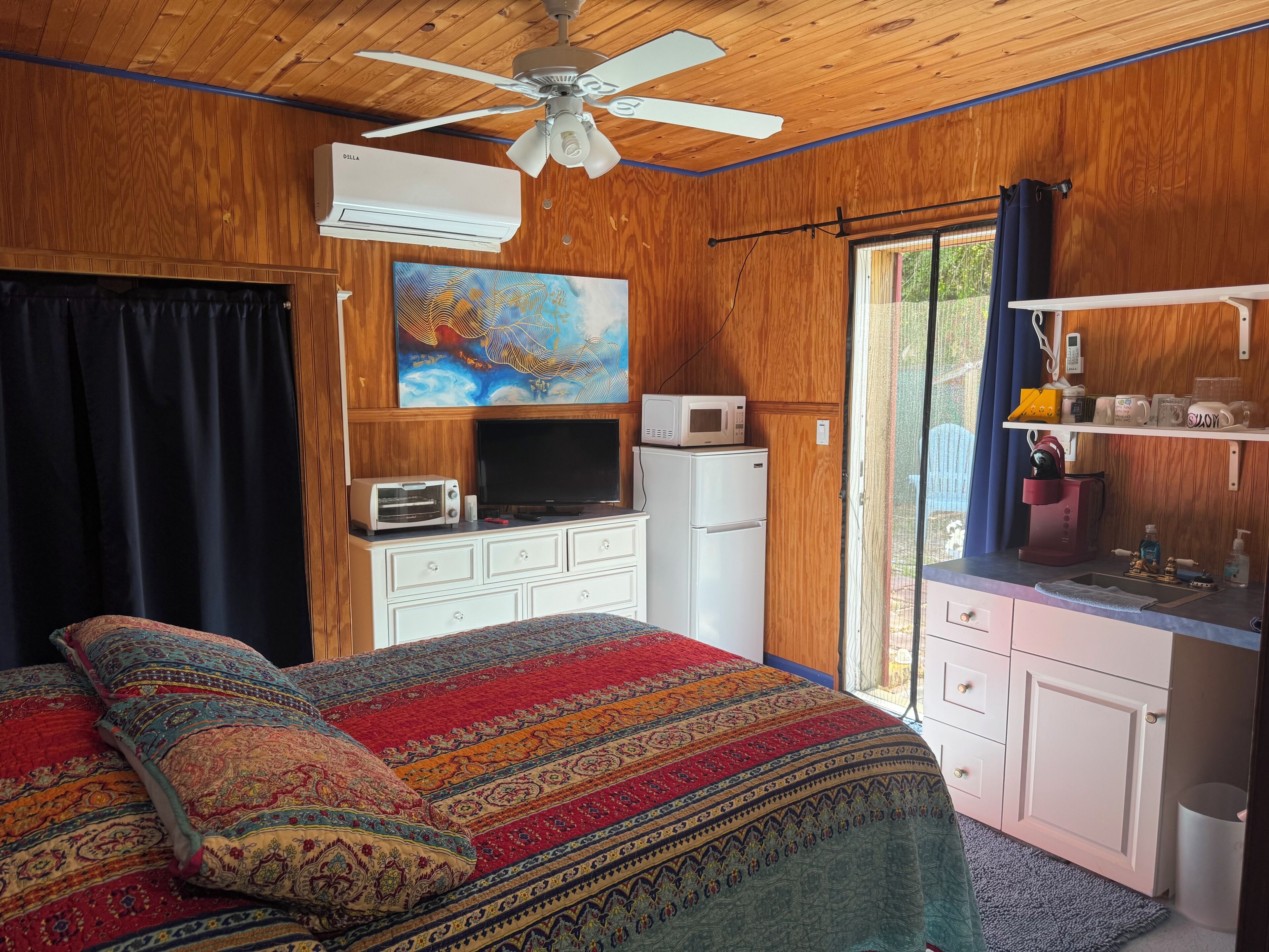 A bedroom with a bed, a ceiling fan, an air conditioner, a microwave, a refrigerator, a TV, and a window with curtains.