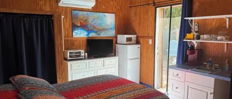 A bedroom with a bed, a ceiling fan, an air conditioner, a microwave, a refrigerator, a TV, and a window with curtains.