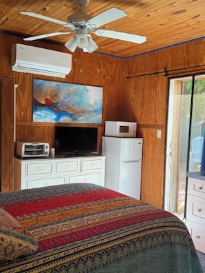 A bedroom with a ceiling fan, an air conditioner, a microwave, a television, a dresser, and a patterned bedspread.