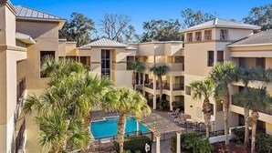 Un hotel de varios pisos con un patio central que cuenta con una piscina, rodeado de palmeras y sillas de descanso.