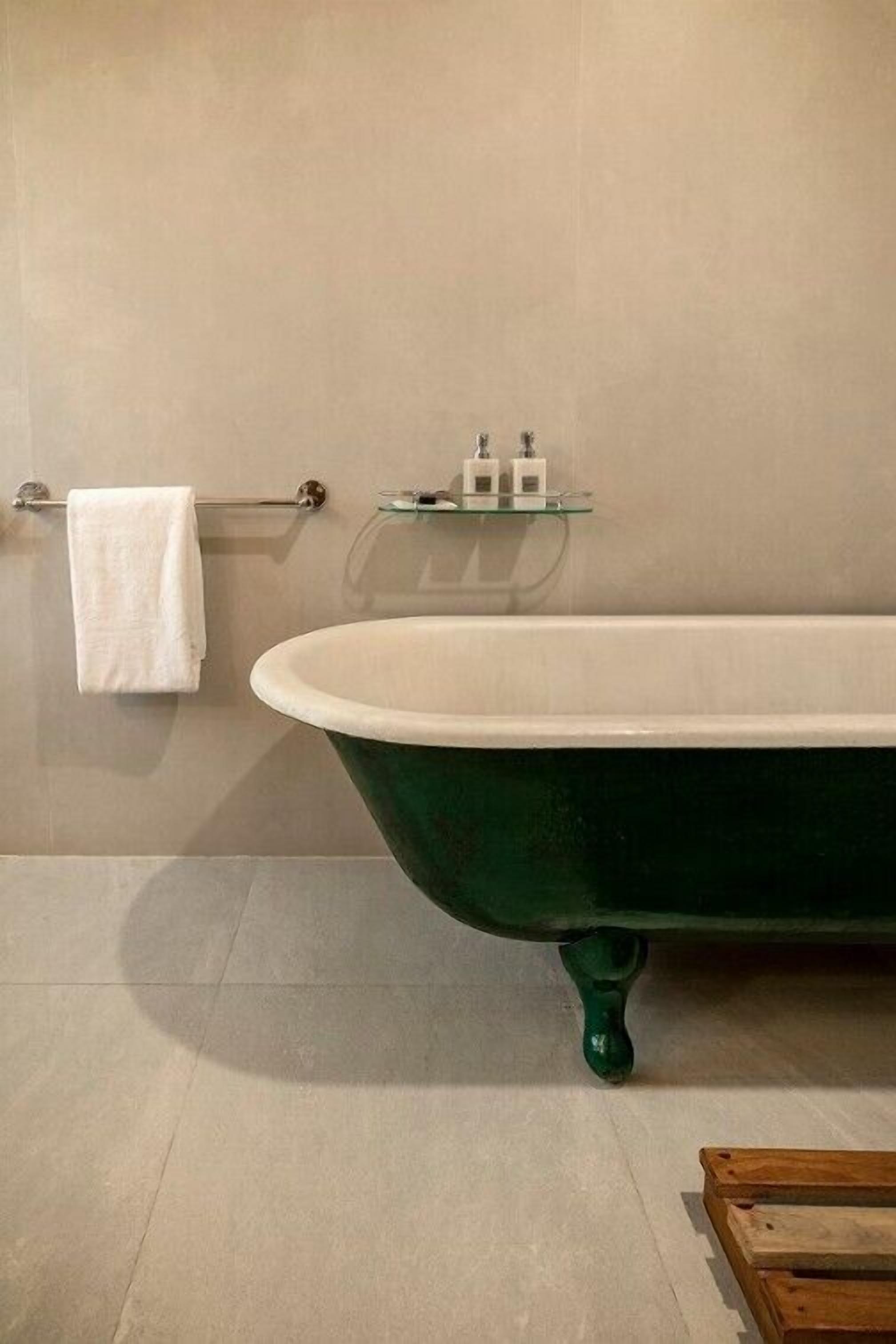 Un baño con una bañera de patas de león verde, una toalla blanca colgada en un perchero y un estante de vidrio con artículos de tocador.