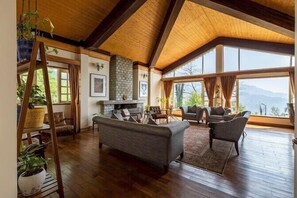 A spacious living room with a high ceiling, wooden beams, a fireplace, and large windows offering a view of the outdoors. - Amã Stays & Trails Makaibari Bungalow, Darjeeling (Kurseong)