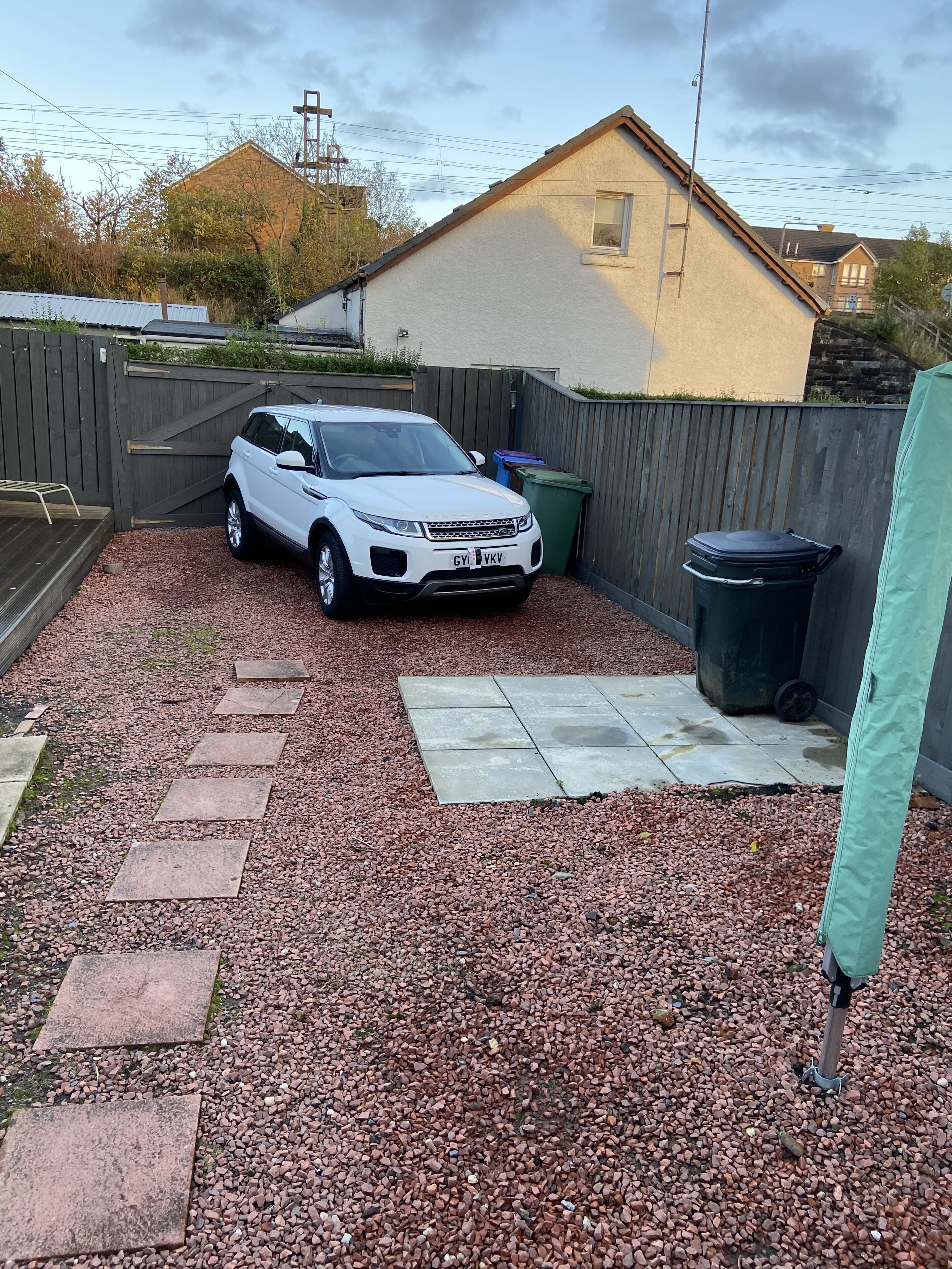En vit SUV står parkerad på en grusuppfart, med ett trästaket och ett hus i bakgrunden.