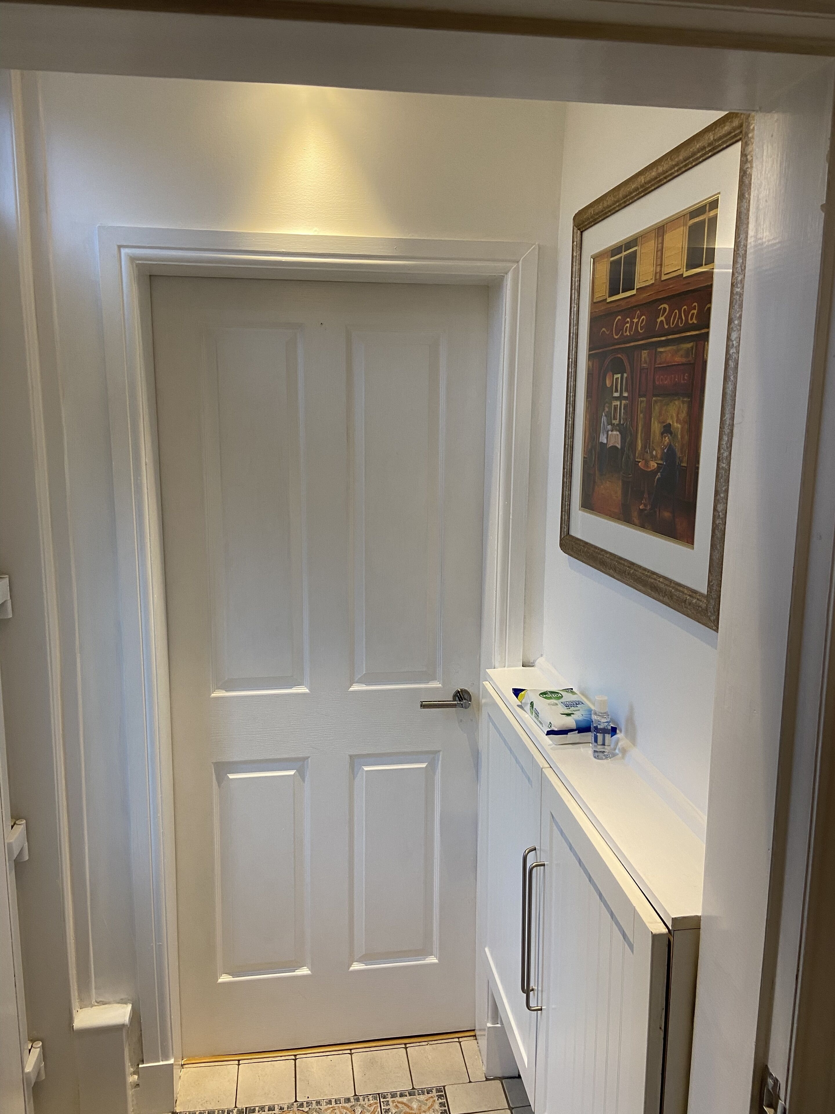 Een witte deur met een deurklink, een betegelde vloer, een klein plankje met een ingelijst foto en een witte muur.