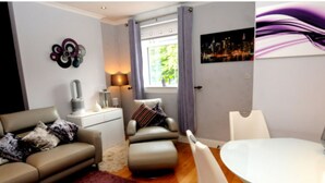 Ein modernes Wohnzimmer mit einer Couch, einem Hocker und einem Esstisch mit Stühlen. Ein Fenster mit Vorhängen, eine Lampe und ein Stadtbild an der Wand sind ebenfalls vorhanden.