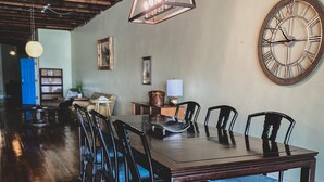 Un espace repas avec une longue table en bois, des chaises noires dotées de coussins bleus, une grande horloge murale et un luminaire suspendu.