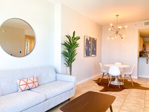 Una sala de estar moderna con un sofá azul claro, un espejo redondo, una planta en maceta, una zona de comedor con una mesa y sillas blancas, y una araña.