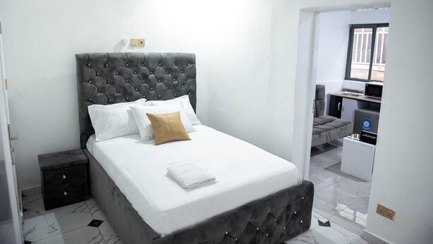 A modern bedroom with a tufted headboard, a neatly made bed with white linens and a brown pillow, a small bedside table, and a desk with a computer and printer. - Lovely One-Bed Apartment Located in Abuja (Abuja)
