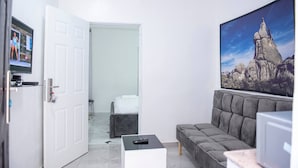 A modern room with a grey sofa, a small white table, a remote control, a door leading to a bathroom, and a large rock-themed painting on the wall. - Lovely One-Bed Apartment Located in Abuja (Abuja)