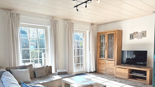 A living room with a sofa, a coffee table, a TV on a wooden stand, and large windows with curtains. - Holiday Home by the Lake with Garden and Terrace (Haselünne)