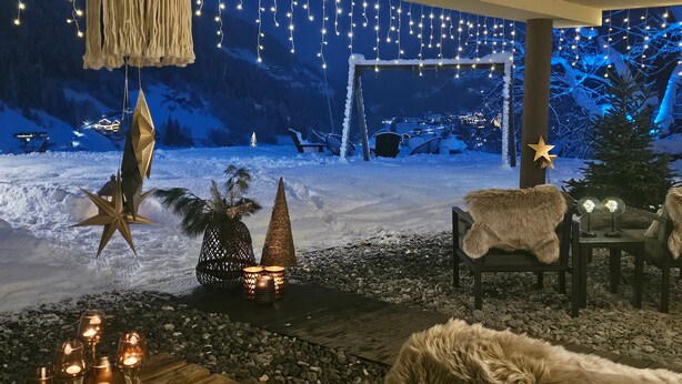 Ein Sitzbereich im Freien mit verschneiter Landschaft, Lichterketten und dekorativen Elementen wie einem Stern und einer kegelförmigen Struktur.