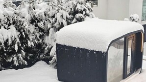 Ein mit Schnee bedeckter Außenbereich mit einer dunkel gefärbten Struktur, umgeben von schneebedeckten Bäumen und einem Gebäude im Hintergrund.