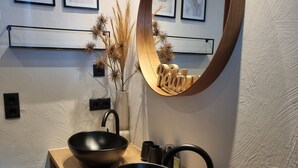 Ein Badezimmer mit zwei schwarzen Waschbecken, einem Holzkosmetikschrank, einem runden Spiegel und gerahmten Motivationssprüchen an der Wand.