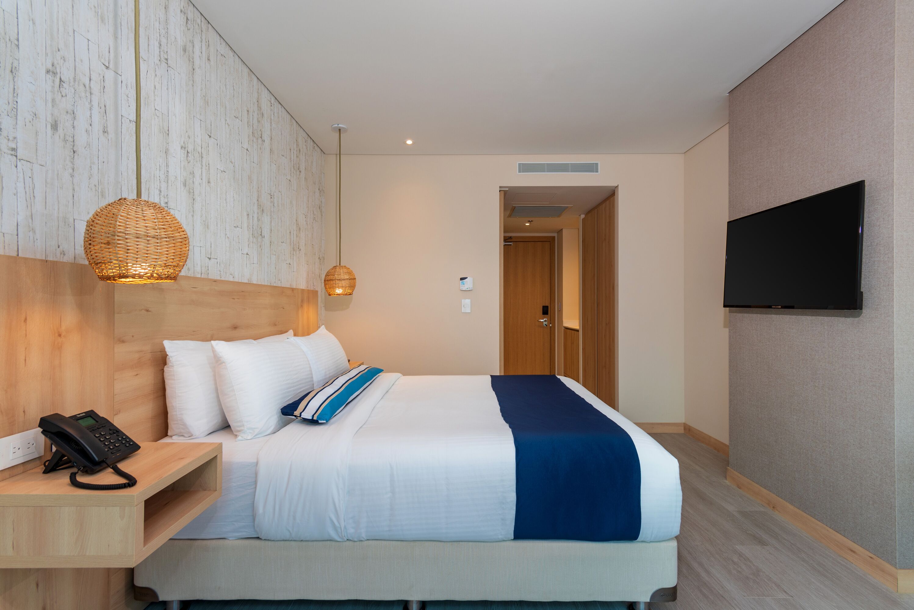 Quarto de hotel moderno com cabeceira de madeira, cama com roupa de cama branca e azul, telefone ao lado da cama, luminária pendente de fibra e televisão de tela plana na parede.