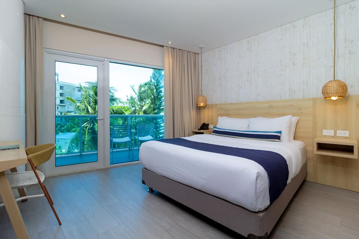 Une chambre d’hôtel avec un grand lit, un bureau avec une chaise, un balcon donnant sur des palmiers et une tête de lit en bois.