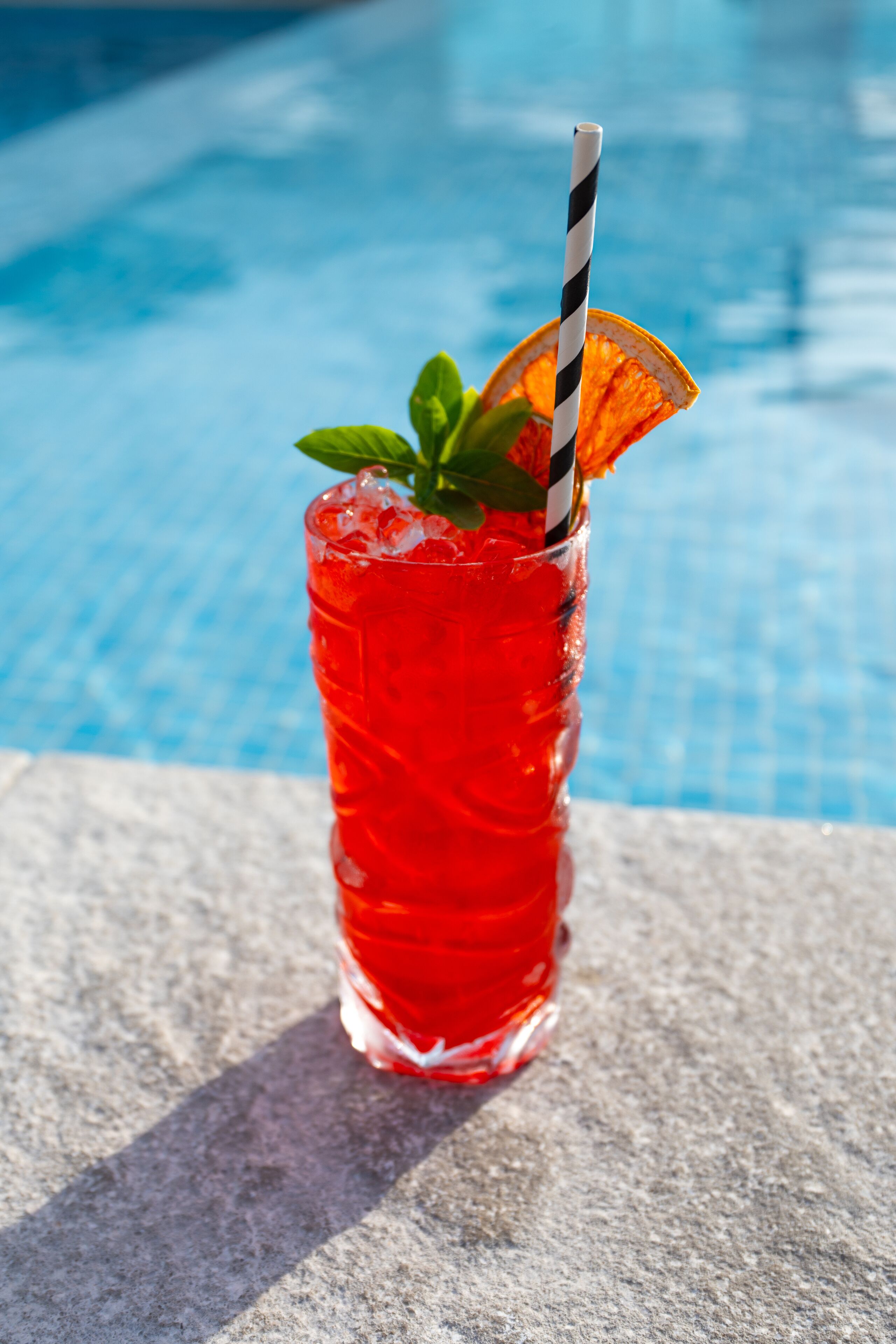 A glass of red beverage with a striped straw and an orange slice garnish, placed on a stone surface by a pool.