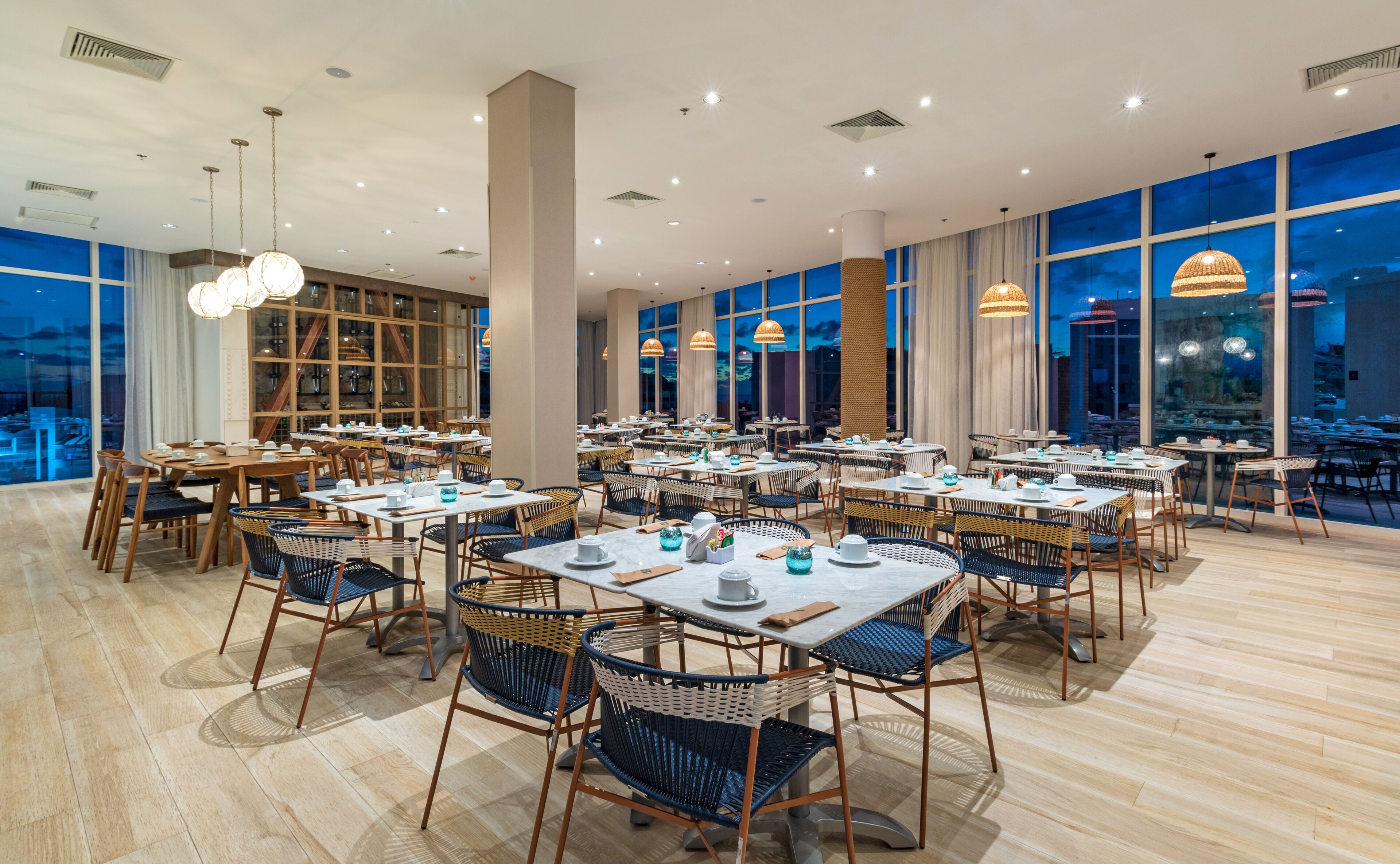 A spacious dining area with multiple tables and chairs, large windows offering views of the city, and modern lighting fixtures.