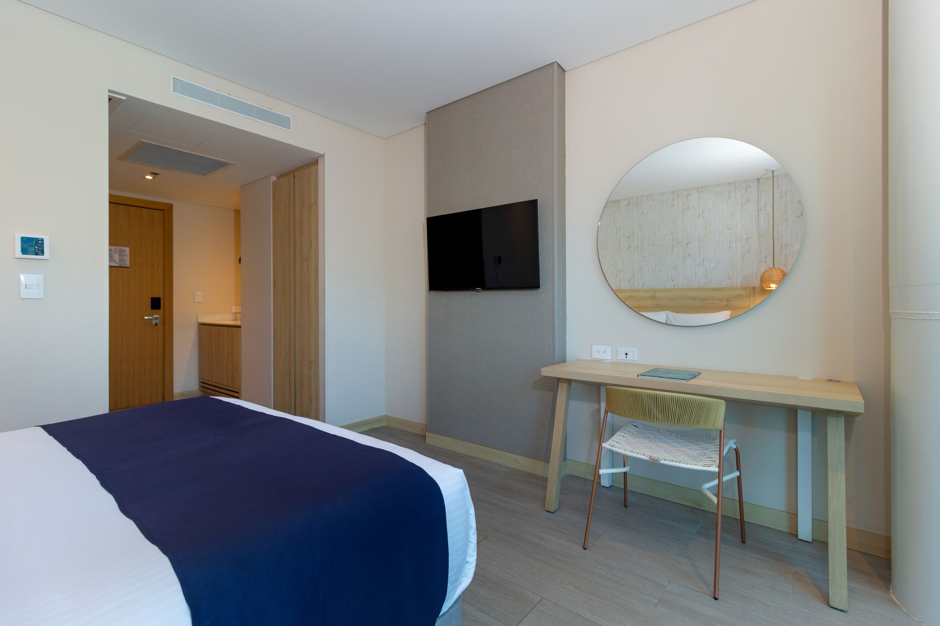 A hotel room with a bed, a desk, a chair, a television, and a round mirror.