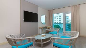 A modern hotel room with a round glass table, wicker chairs with blue cushions, a flat-screen TV mounted on the wall, and a large window offering a view of the city.