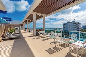 A rooftop terrace with lounge chairs, a pool, and a view of the ocean and buildings.