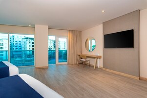 A hotel room with a large bed, a desk, a chair, a TV, and a view of the city through the window.